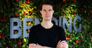 A man with short dark hair, wearing a black t-shirt, stands with arms crossed in front of a green leafy wall with the word "BENDING" partially visible behind him.