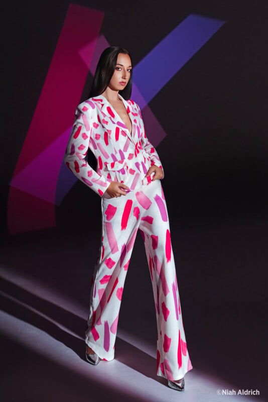 A woman poses confidently in a white suit with bold pink and red patterns, standing against a dark background with abstract purple and red geometric shapes. She has long, straight hair and is wearing silver heels.