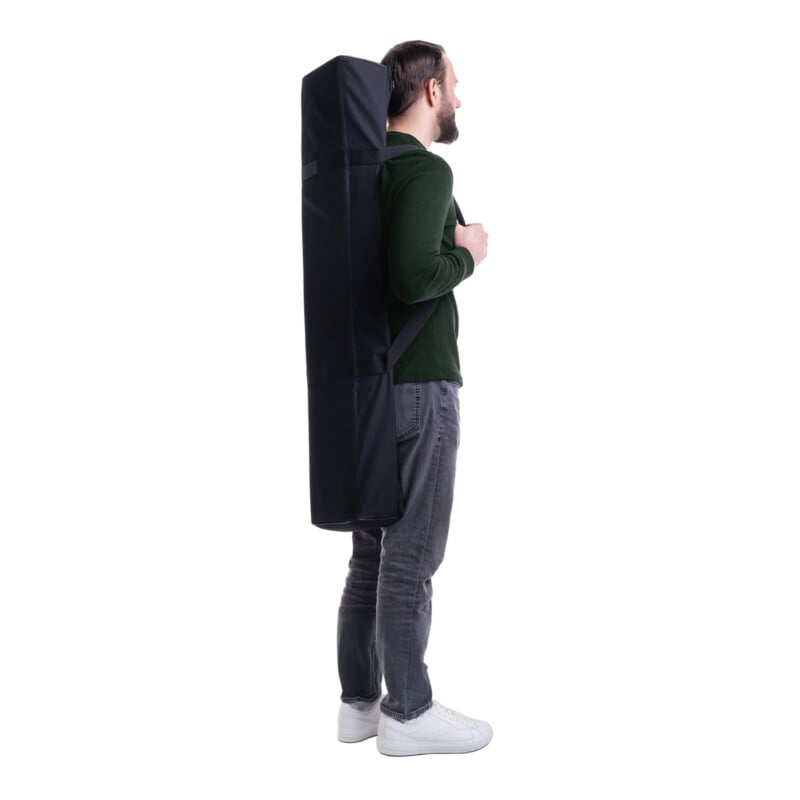 A man in a green sweater and gray jeans stands in profile, carrying a large black rectangular bag on his back with a shoulder strap, against a white background.