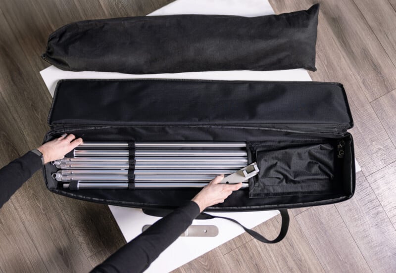 A person’s hands are packing or unpacking metal rods and a tool from a black carrying case on a wooden floor, with another black bag and white rectangular surfaces nearby.