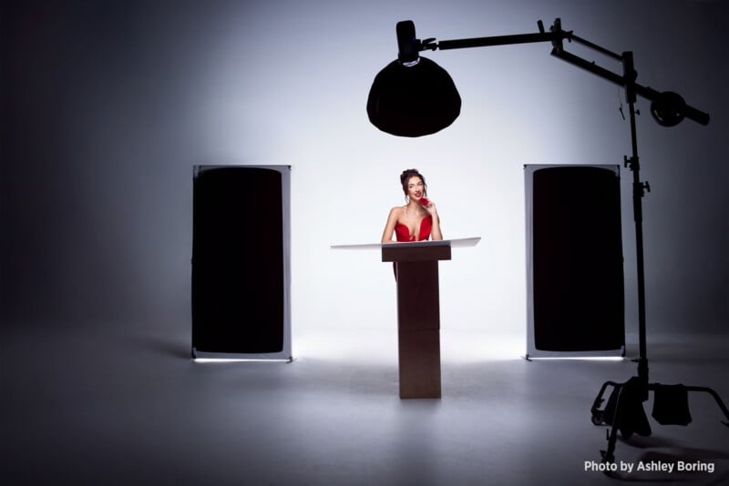 A woman in a red dress stands at a podium in a photo studio, surrounded by lights and black panels, with photo equipment visible around her. The background is white and well-lit. Photo by Ashley Boring.