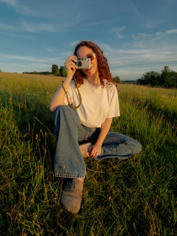 A person with curly hair wearing sunglasses, a white t-shirt, and blue jeans sits on grass in a sunlit field, holding a camera up to their face as if about to take a photo.