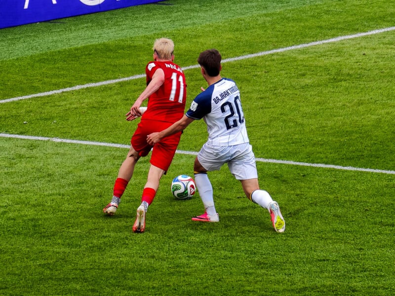 A soccer player in a red uniform tries to control the ball while being closely defended by an opponent in a white uniform during a match on a green grass field.