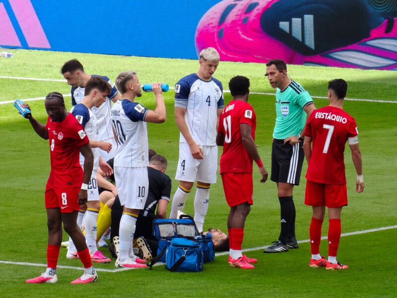 Soccer players from two teams and a referee stand around a player lying on the field receiving medical attention, while other players drink water and talk during a break in the match.