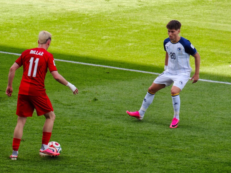 A soccer player in a red uniform, number 11, faces an opponent in a white and blue uniform, number 20, on a green soccer field during a match.
