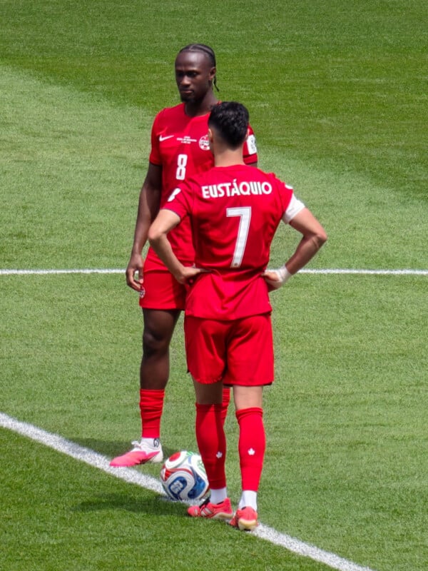 Two soccer players in red Canada uniforms stand on a green field during a match. One player faces forward with hands behind back, while the other, seen from behind, stands with hands on hips near the ball.