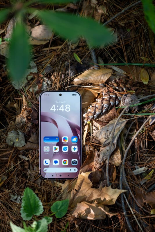 A smartphone with a lit screen showing the home screen lies on the forest floor among dry leaves, pine needles, and a pine cone, partially shaded by green foliage.