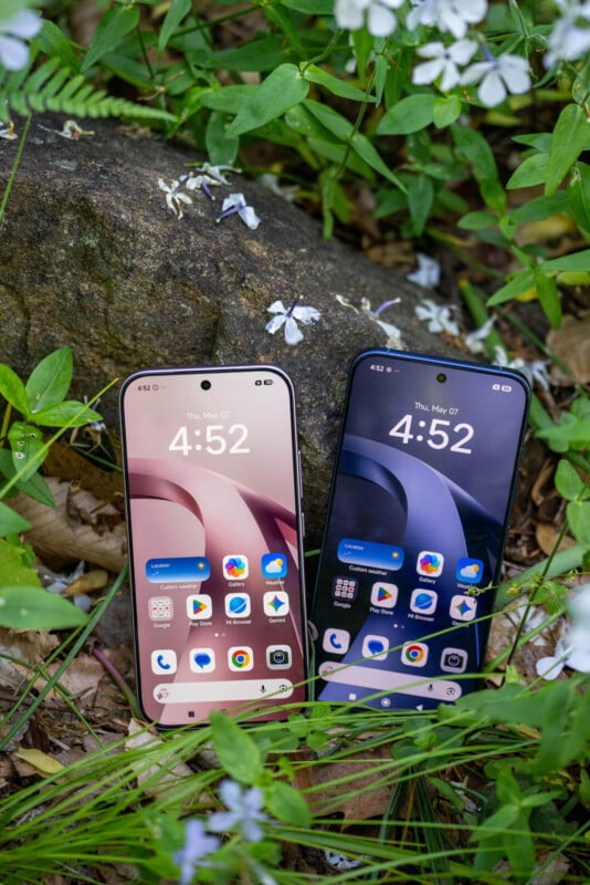 Two smartphones with different wallpaper colors, one pink and one blue, are standing upright side by side on the ground among green plants, flowers, and rocks. Both screens display the time 4:52 and similar app icons.