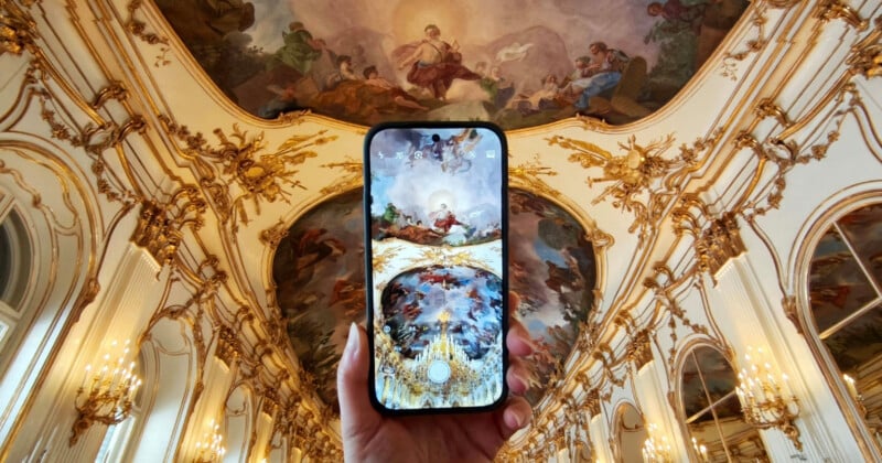 A hand holds up a smartphone displaying a digital version of the ornate, painted ceiling above in a grand hall decorated with gold accents, chandeliers, and intricate frescoes.