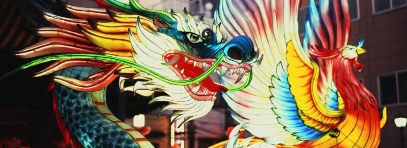A vibrant, illuminated dragon and phoenix lantern display with vivid colors and detailed patterns, set against an urban backdrop at night.