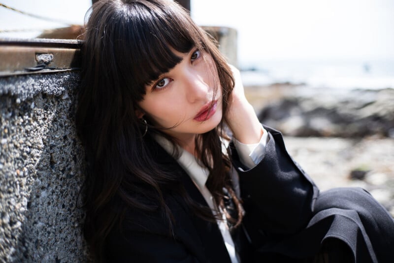 A woman with long dark hair and bangs, wearing a black blazer and white shirt, sits by a rough concrete wall outdoors, touching her hair and gazing thoughtfully at the camera. The background is bright and blurred.