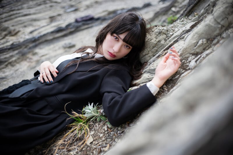 A woman with long dark hair and bangs, dressed in a black coat, lies on rocky ground, leaning against a large rock. She looks at the camera with a serious expression. Some small plants grow near her hand.