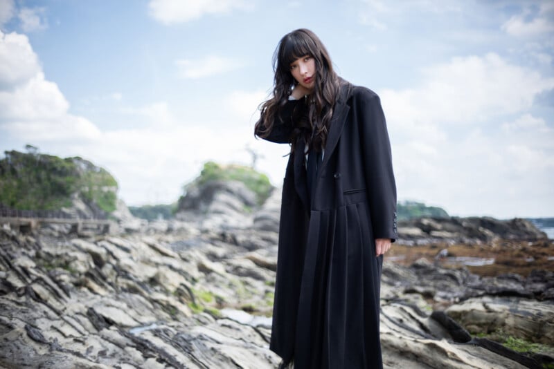 A person with long dark hair wearing a long black coat stands on rocky terrain by the seaside, with a cloudy blue sky and distant greenery in the background.