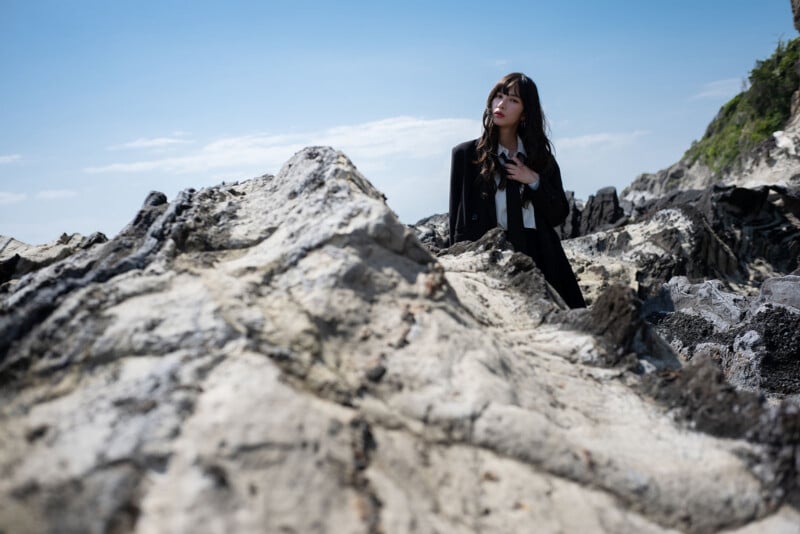A woman with long dark hair, wearing a black coat, stands among large rocky formations under a blue sky with scattered clouds. She looks thoughtfully into the distance.