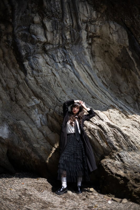 A woman in dark, layered clothing and sunglasses poses against a large, textured rock formation, standing on a rocky surface with sunlight highlighting parts of the scene.
