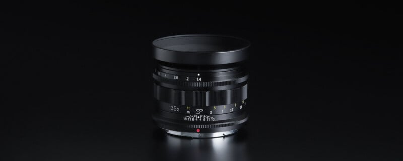 A black camera lens with white and yellow markings is standing upright on a smooth, dark surface, set against a black background.