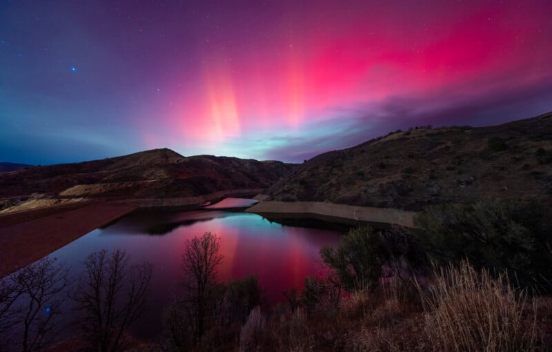 Bright pink and purple aurora lights shine in the night sky over a calm reservoir, reflecting on the water, with hills and sparse vegetation in the foreground.