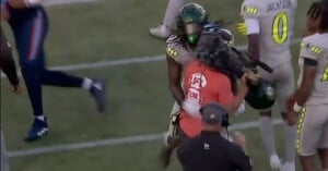 A football player in a gray and green uniform collides with a camera operator wearing an orange FOX shirt on the sidelines during a game. Other players are nearby on the field.