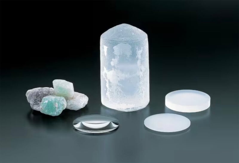 Several rough green and white crystals, a large clear synthetic crystal, and three polished optical lenses are displayed on a dark surface, illustrating the process of transforming raw minerals into finished lenses.