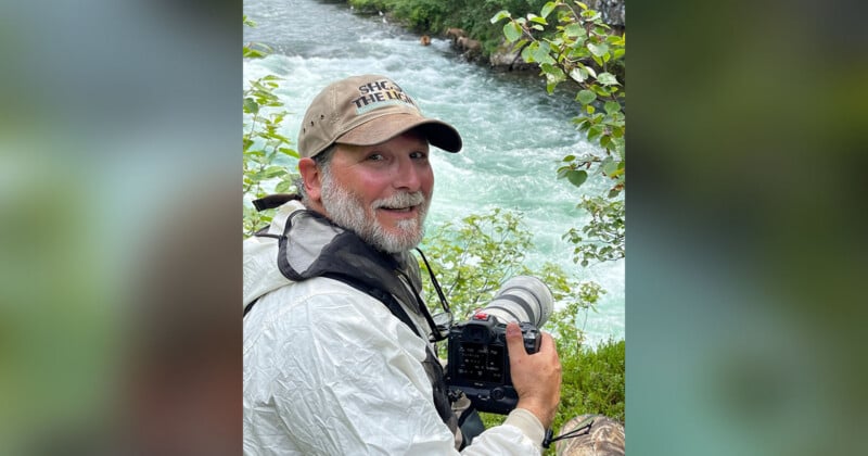 A bearded man in outdoor gear and a cap smiles at the camera while holding a professional camera by a fast-flowing river, surrounded by green foliage.