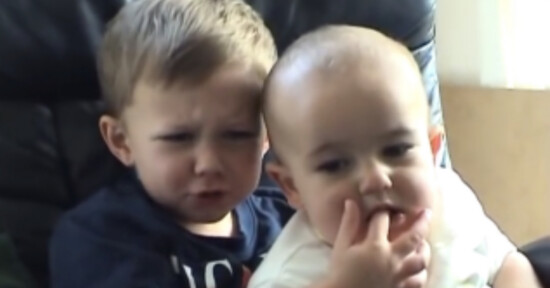 Two young children sit close together. The older child looks upset while the younger child places a finger in the older child’s mouth, causing a funny and slightly distressed expression.