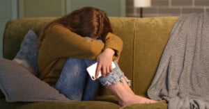 A person with long hair sits on a green couch, hugging their knees and resting their head on their arms. They are holding a smartphone and appear distressed. A gray blanket is draped on the couch beside them.