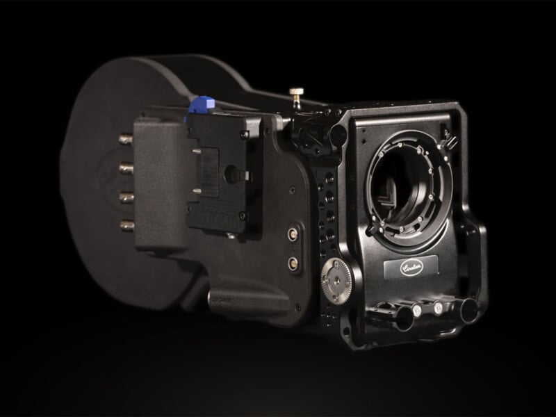 A black, professional film camera body with a large round film magazine attached, various control dials, and an empty lens mount, displayed against a black background.