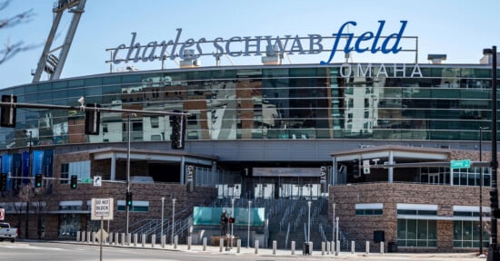 Exterior view of Charles Schwab Field Omaha, showing large signage on the stadium, glass windows, entrance stairs, and surrounding city buildings on a clear day.