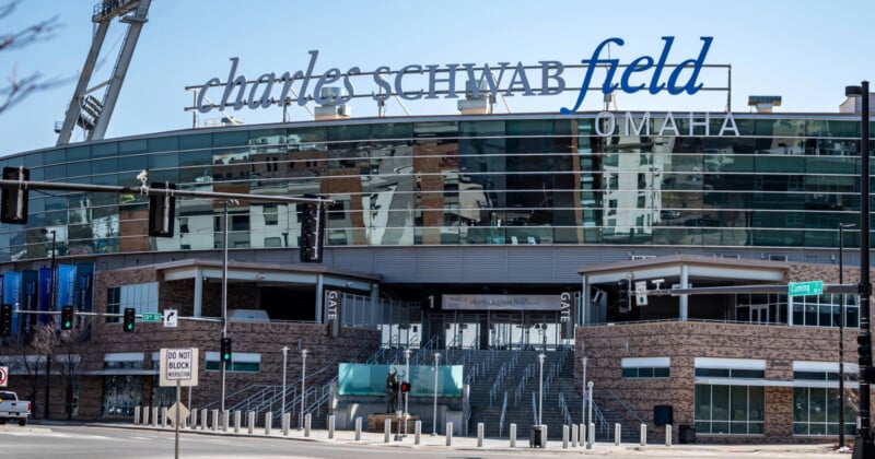 Exterior view of Charles Schwab Field Omaha, showing large signage on the stadium, glass windows, entrance stairs, and surrounding city buildings on a clear day.