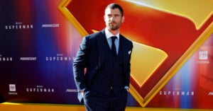 A man in a dark pinstripe suit stands with his hands in his pockets in front of a large Superman logo backdrop at a red carpet event.