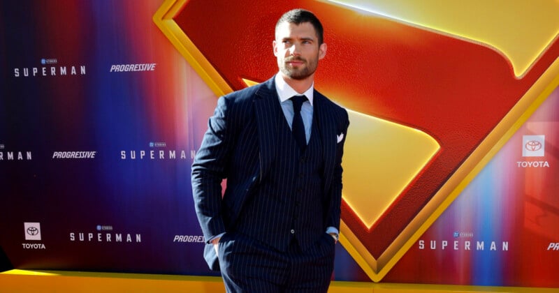 A man in a dark pinstripe suit stands with his hands in his pockets in front of a large Superman logo backdrop at a red carpet event.