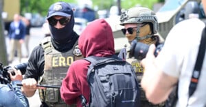 Two law enforcement officers, one in a helmet and one in a cap with "ERO" on a vest, confront a person in a red hoodie and black backpack while photographers capture the scene outdoors.