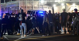 A large group of people gathers on a street at night near police vehicles with flashing lights; some people are taking photos or videos, and a high fence is visible in the background.