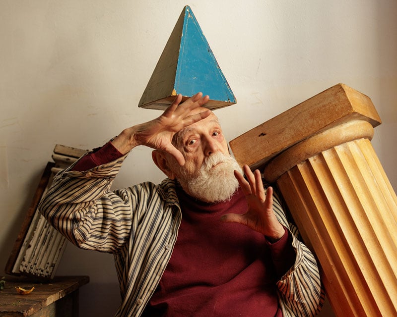 Un anciano con barba blanca se sienta en el interior y lleva un pilar triangular azul en la cabeza. Hizo un gesto con las manos y se apoyó contra un gran pilar caído. Llevaba una camisa a rayas y un top granate.