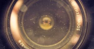 Close-up view of a camera lens with visible dust particles on the glass surface. The lens rings and some text around the edges are also visible.