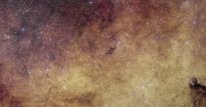 A dense field of stars and cosmic dust fills the image, creating a golden and reddish nebula-like effect, with bright and faint stars scattered throughout the scene.