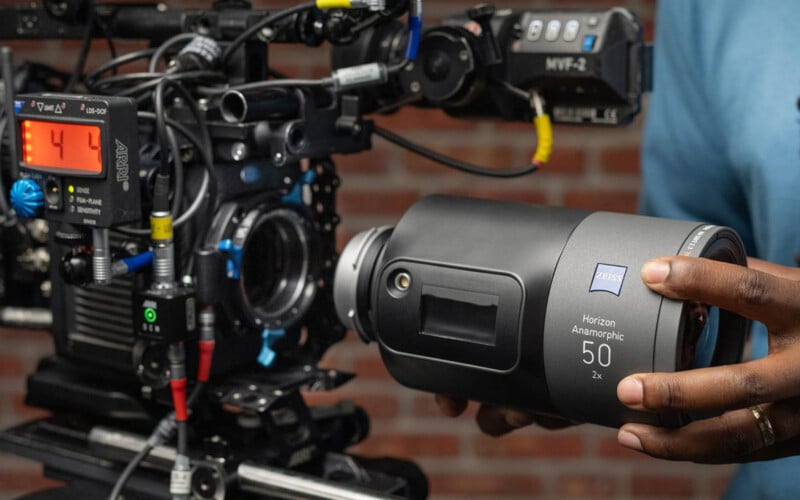 A person holds a ZEISS 50mm Horizon Anamorphic lens next to a professional cinema camera with a digital display and cables, against a blurred brick wall background.