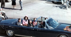 A motorcade with four people in an open-top car; a man and woman sit in the backseat while another man and driver are in front. The car has an American flag and is surrounded by people, motorcycles, and other vehicles. jfk assasination john f kennedy