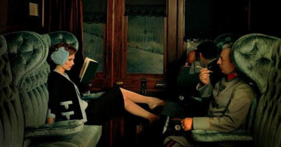 A woman in earmuffs lounges with her legs on a train seat, reading a book, while two men in vintage military uniforms sit across from her, one smoking and the other looking out the window.