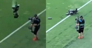 A camera operator stands on a football field as a large camera drone crashes to the ground nearby, breaking into pieces. The operator is holding a camera and wearing athletic gear.