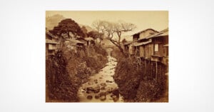 Sepia-toned photo of a narrow rocky stream flowing between traditional wooden houses on stilts, with bare trees and mountains visible in the background.