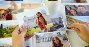 A person holds a photo of a smiling woman in front of a historic building while arranging several printed travel photos on a table, with a mug and more photos visible in the background.