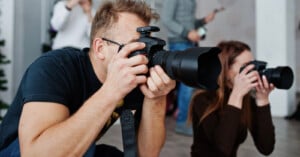 Two people crouch while holding cameras, aiming their lenses forward to take photographs. Other blurred figures are visible in the background, suggesting a group setting or event.