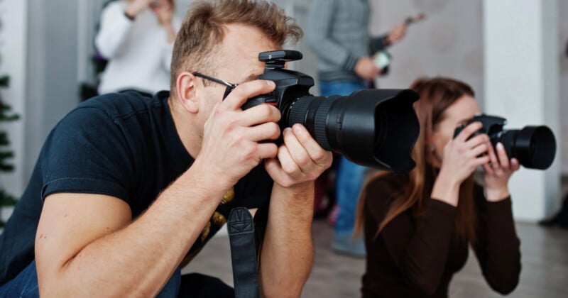 Dos personas estaban en cuclillas, sosteniendo cámaras y apuntando la lente frente a ellos para tomar fotografías. Se pueden ver otras figuras borrosas en el fondo, lo que sugiere que se trata de un evento o escenario de grupo.
