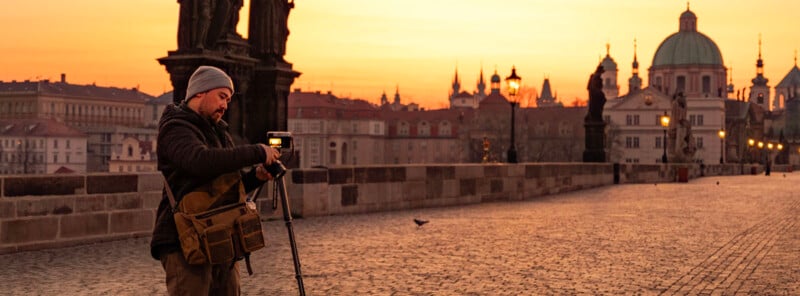 A man adjusts a camera on a tripod at sunrise on a cobblestone bridge, with historic buildings and domes in the background, under an orange sky.