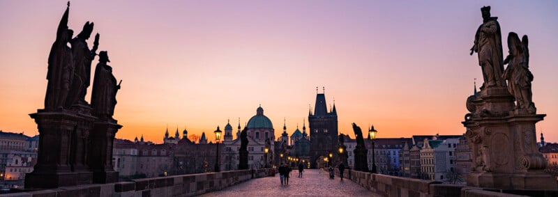 People walk along the historic Charles Bridge in Prague at sunset, with statues lining the bridge and the city skyline, including church domes and towers, silhouetted against a colorful sky.