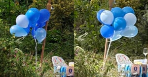 A bunch of blue and light blue balloons is tied to a stick outdoors amid greenery. Nearby are chairs, a baby-themed block, and a wine glass, suggesting a party or celebration in a garden setting.