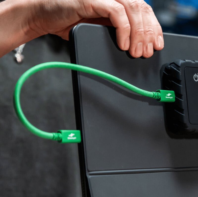 A hand holds a device connected by a green charging cable to a black power bank, with both cable ends featuring green labels marked "Step Up.
