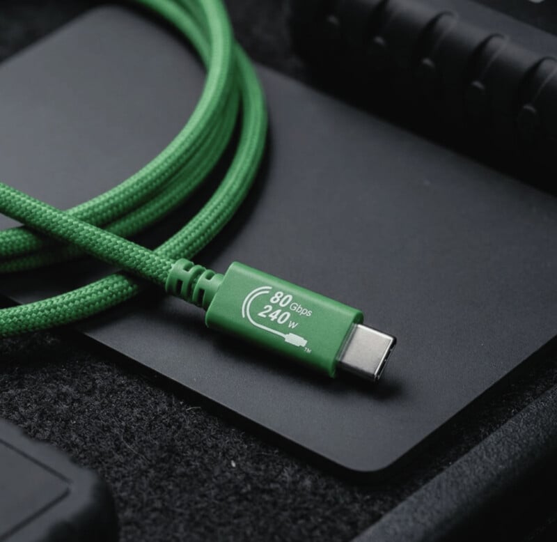 A green braided USB-C cable labeled "80 Gbps 240 W" lies on a dark surface next to electronic accessories.