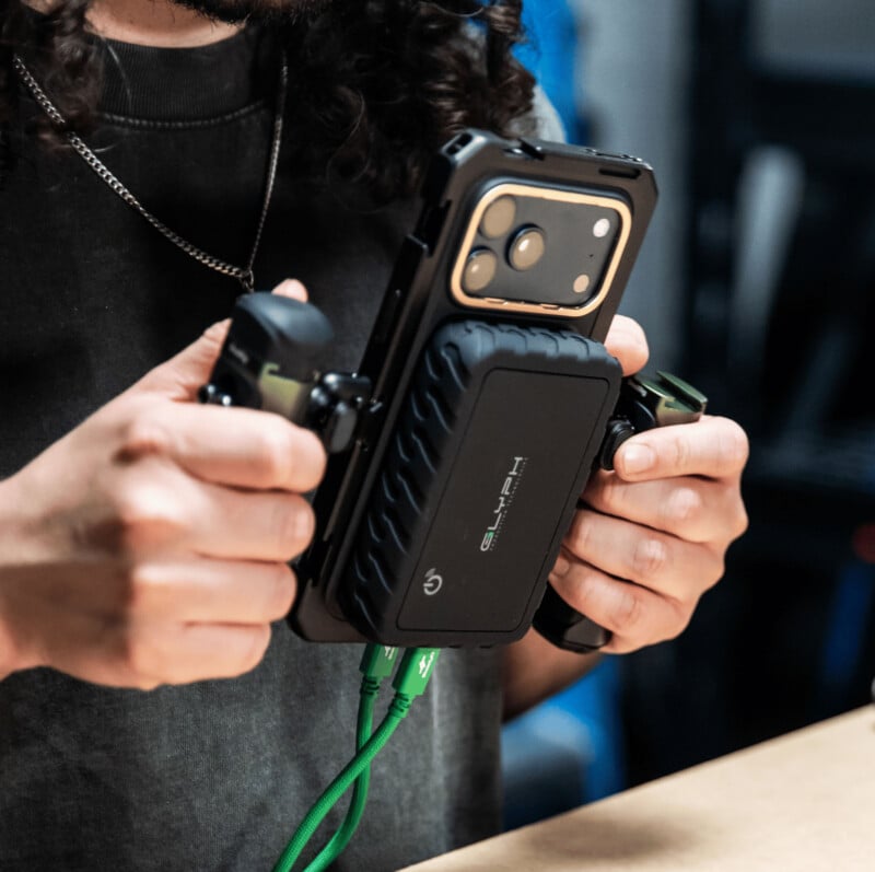 A person holds a smartphone mounted in a rugged gaming controller accessory, with green cables connected at the bottom. The background is blurred, focusing on the hands and device.